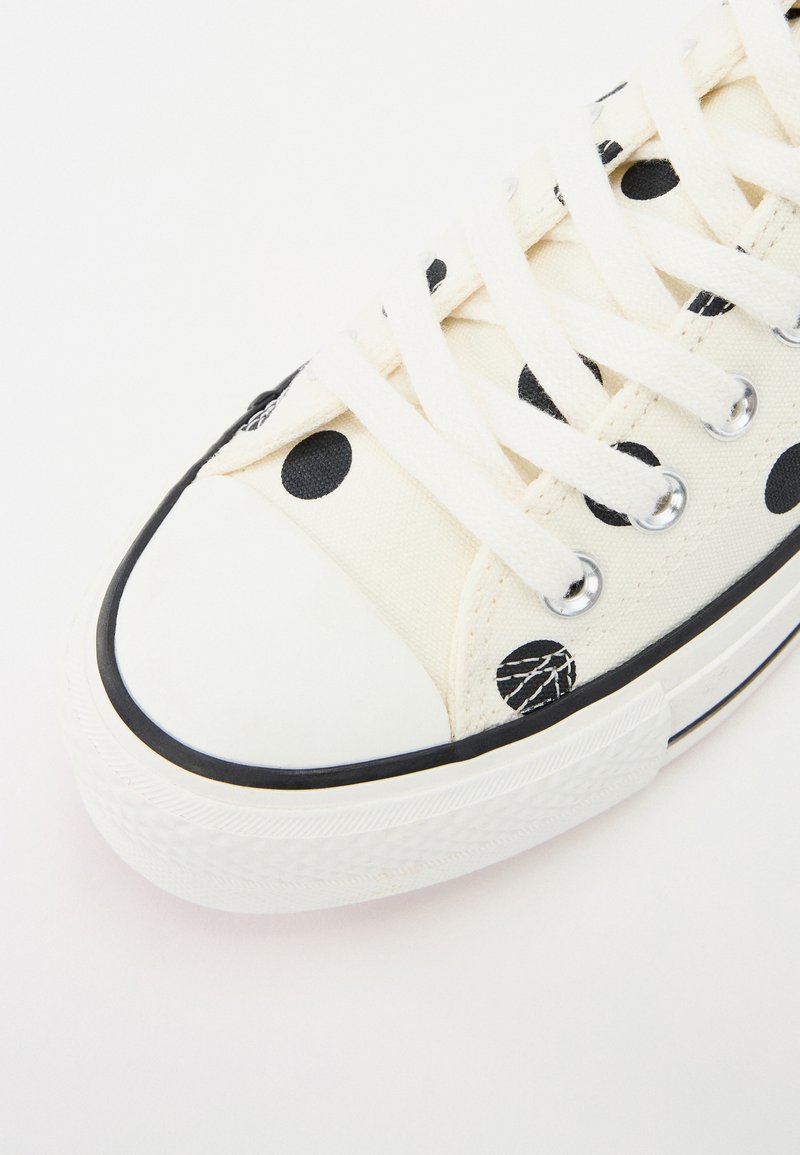 Zapatilla de lona con lunares negros, puntera blanca, suela de goma negra y cordones blancos. Cuenta con ojales metálicos y un tejido texturizado.