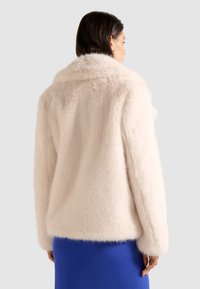 Faux fur jakke i blød blush pink, der har en afslappet pasform, bred krave og lange ærmer, båret over en blå nederdel.