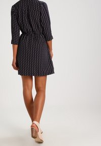 Robe marine avec une taille cintrée, des manches 3/4 et un motif blanc. Le tissu semble léger, associé à des sandales à talons beiges.