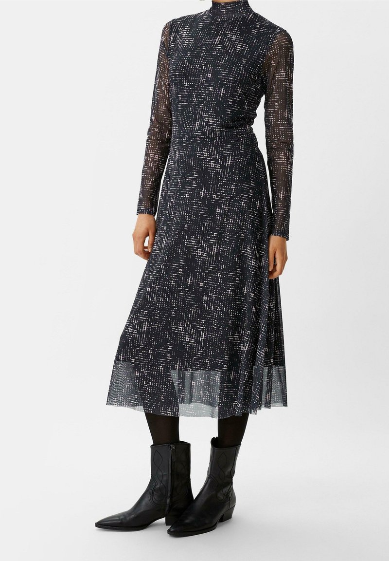 Langärmliges schwarzes Kleid mit einem strukturierten weißen Muster, mit hohem Kragen und einem fließenden Rock. Inklusive schwarzer Ankle Boots mit spitzen Zehen.