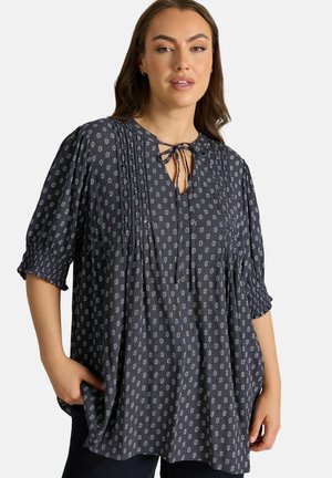 Frau trägt eine locker sitzende dunkelblaue Bluse mit kleinen weißen Blumenmustern, Biesen-Details, Dreiviertelärmeln und einer Kordel am Halsausschnitt.