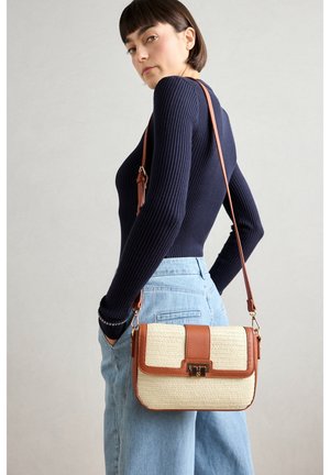 Femme portant un pull côtelé bleu marine et un jean bleu clair, avec un sac bandoulière en cuir beige et marron sur l'épaule.