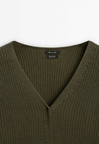 Maglione lavorato a maglia verde oliva con scollo a V, realizzato in filato di cotone spesso, che mostra una trama di punti dettagliata e l'etichetta del marchio sul collo.