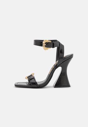 Sandale à talon haut en cuir verni noir avec un talon bloc évasé unique, une bride à la cheville et des boucles décoratives dorées sur les brides des orteils et de la cheville.