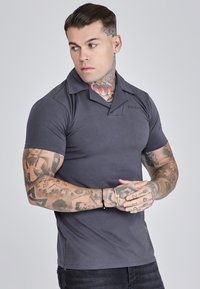 Polo gris de manga corta con un cuello profundo, hecho de una tela suave. Presenta un sutil detalle de logo en el pecho. Diseño ajustado.