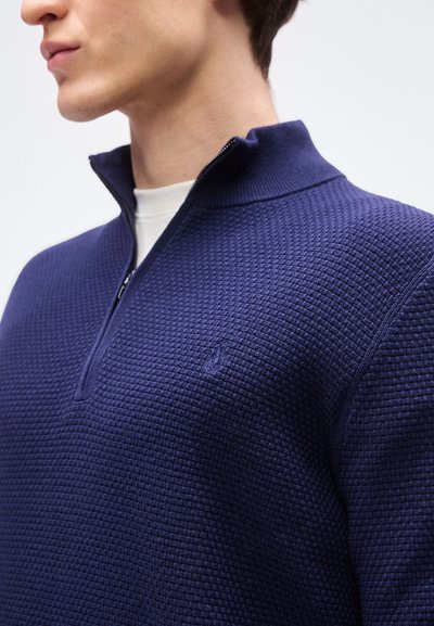 Νέος άνδρας που φοράει μπλε ναυτικό πουλόβερ με υφή και φερμουάρ μέχρι το μέσο του λαιμού πάνω από ένα λευκό πουκάμισο, εμφανίζεται από τους ώμους και πάνω σε κοντινή πλάγια όψη.