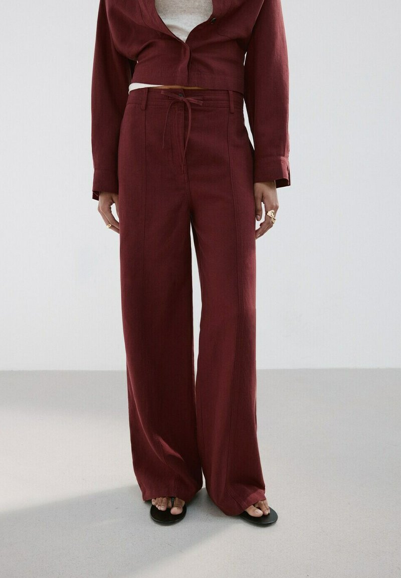 Femme portant un pantalon bordeaux taille haute à jambes larges avec une taille à cordon, assorti d'une veste courte assortie et de sandales noires.