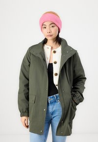 Veste imperméable vert olive avec un grand col, des boutons-pression et des poches latérales, portée sur un pull côtelé blanc et un jean bleu, avec un bonnet rose.