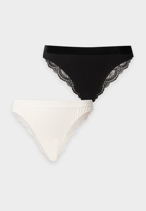 Femme aux longs cheveux foncés portant un soutien-gorge en dentelle blanche et un sous-vêtement blanc assorti, debout pieds nus sur un fond blanc uni.