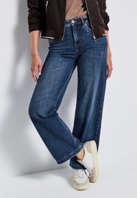 Jean large en denim bleu foncé avec une texture lisse, présentant une fermeture éclair et un bouton, associés à des baskets blanches.
