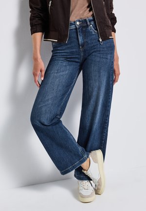 Brede jeans in donkerblauwe denim met een gladde textuur, voorzien van een ritssluiting en knoopsluiting, gecombineerd met witte sneakers.