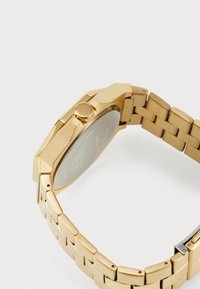 Guldmetallarmbandsur med en oktagonal klockkåpa, utrustat med ett texturerat armband, rund krona och en slät, reflekterande baksida.