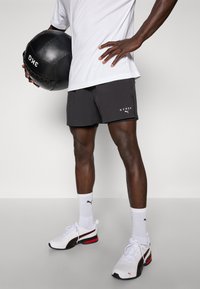Shorts de sport noirs avec un logo blanc, associés à un t-shirt blanc et des baskets blanches avec des accents rouges et noirs, tenant un ballon de médecine noir.
