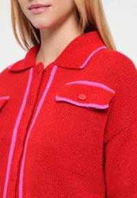 Sarkans adīts kardigans ar rozā akcentiem, lielu apkakli un kabatām ar pogām. Raksturīga mīksta tekstūra un kontrastējošas svītras priekšpusē.