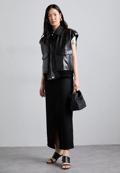 Zwarte leren vest met oversized schouders, twee voorzakken, gecombineerd met een zwarte lange rok en platte zwarte sandalen. Kleine geweven zwarte handtas.