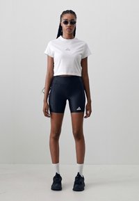 White cropped t-shirt with logo, black fitted shorts, black sneakers, and white socks. Round sunglasses complete the athletic look.