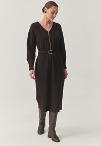 Lange, donkerbruine jurk met een V-hals en een gouden ritssluiting. Heeft een aansluitende riem met een ronde gesp, gecombineerd met kniehoog bruine laarzen.