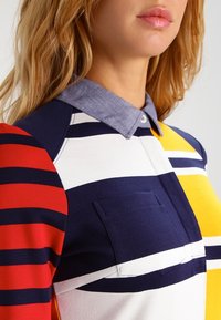 Polo w paski w kolorach czerwonym, granatowym, żółtym i białym; materiał bawełniany o teksturowanej strukturze z kołnierzykiem przypominającym dżins i detalem kieszeni na przodzie.