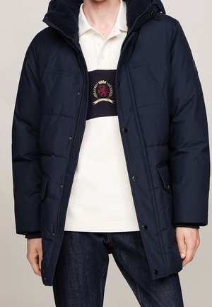 Man wearing a navy blue padded hooded jacket over a white polo shirt with a black horizontal stripe and crest emblem, paired with dark jeans.