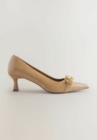 Beige korkokenkä, jossa on terävä kärki ja koristeellinen kultaketju etuosassa, taustalla vaalea tausta.