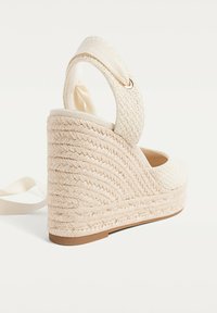 Sandal s potpeticom s pletenim bež gornjim dijelom, s teksturiranom jutenom platformom i mekim tekstilnim remenom oko gležnja za sigurno prianjanje.