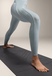 Světlé modré sportovní legíny s texturovanými bočními panely, nošené na černé jógamatce. Noha pevně položená na povrchu jógamatky.