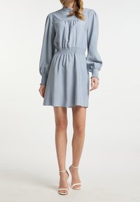 Frau, die ein hellblaues langärmliges Kleid mit gerafftem Bund und Manschetten trägt, kombiniert mit weißen hochhackigen Sandalen mit Knöchelriemen.