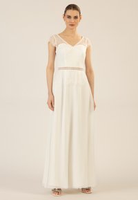 Das weiße Spitzen- und Chiffonkleid hat ein tailliertes Oberteil mit V-Ausschnitt, kurzen Spitzensäumen und einem bestickten Taillenbund, der bis zum Boden fließt.