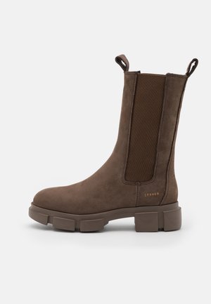 Bottine en daim brun avec des panneaux élastiques sur les côtés, bout arrondi et semelle en caoutchouc épaisse. Elle comporte une languette à l'arrière et un logo de la marque.