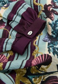 Vícebarevná látka s tyrkysovými, fialovými a zlatými vzory; má řasený rukáv a stříbrný knoflík; měkký, texturovaný materiál.