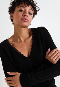 Cardigan noir en tissu doux, avec un col en V orné de dentelle et des manches longues. Il se ferme par des boutons sur le devant.