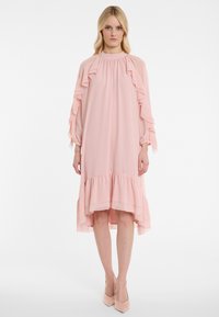 Rosa Chiffonkleid mit Rüschendetails an den Ärmeln und dem Saum, fließendes Design, hoher Kragen und asymmetrischer Saum, kombiniert mit hellrosa Absatzschuhen.