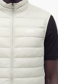 Veste matelassée sans manches gris clair avec fermeture éclair à l’avant et logo EA7 Emporio Armani portée sur un t-shirt noir à manches courtes.