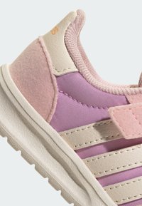 Detailní záběr na růžovou a fialovou dětskou tenisku s béžovým suchým zipem, texturovanou podrážkou a polstrovaným límcem kolem kotníku.
