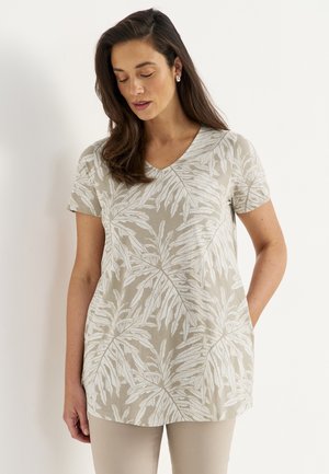 Kvinde iført en beige kortærmet V-hals top med store hvide bladtryk, stående med den ene hånd i lommen, kigger ned.