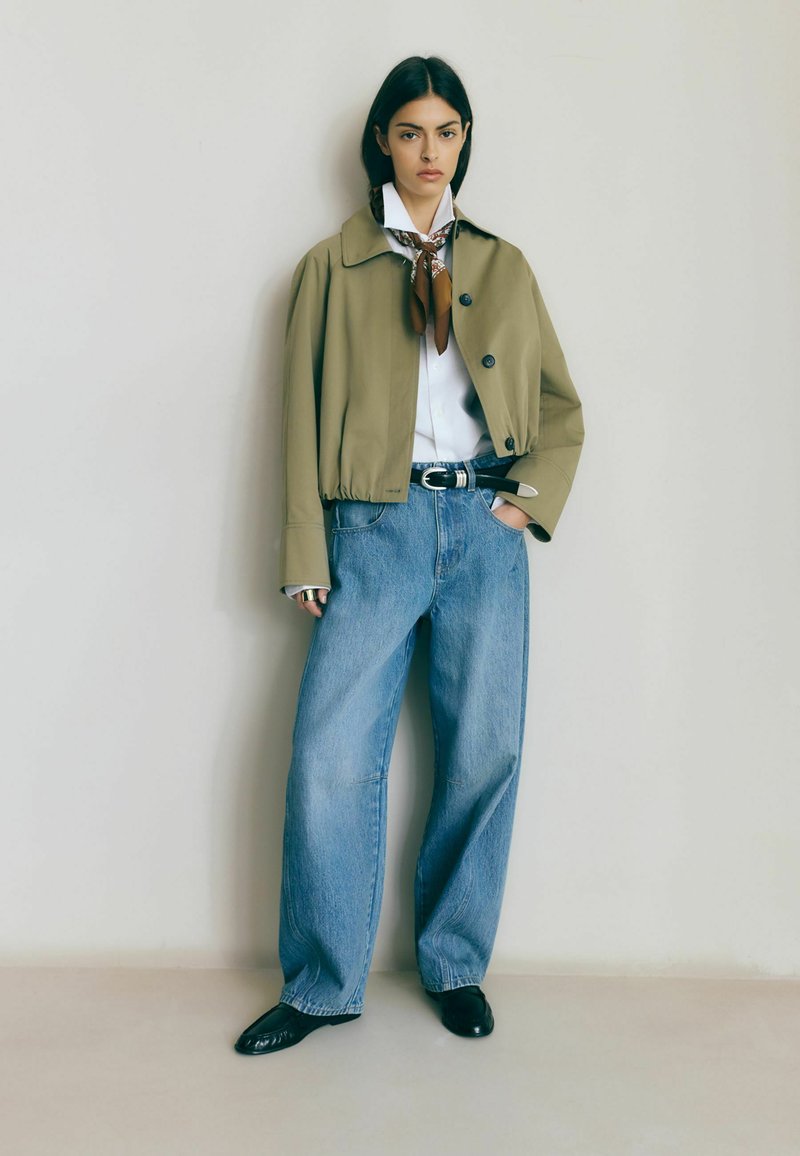 Žena stojaca pri hladkej stene, oblečená v olivovo zelenej skrátenej bunde, bielej košeli s vzorovaným šálom okolo krku, modrých dlhých džínsach s širokými nohavicami, čiernych topánkach a čiernom opasku.