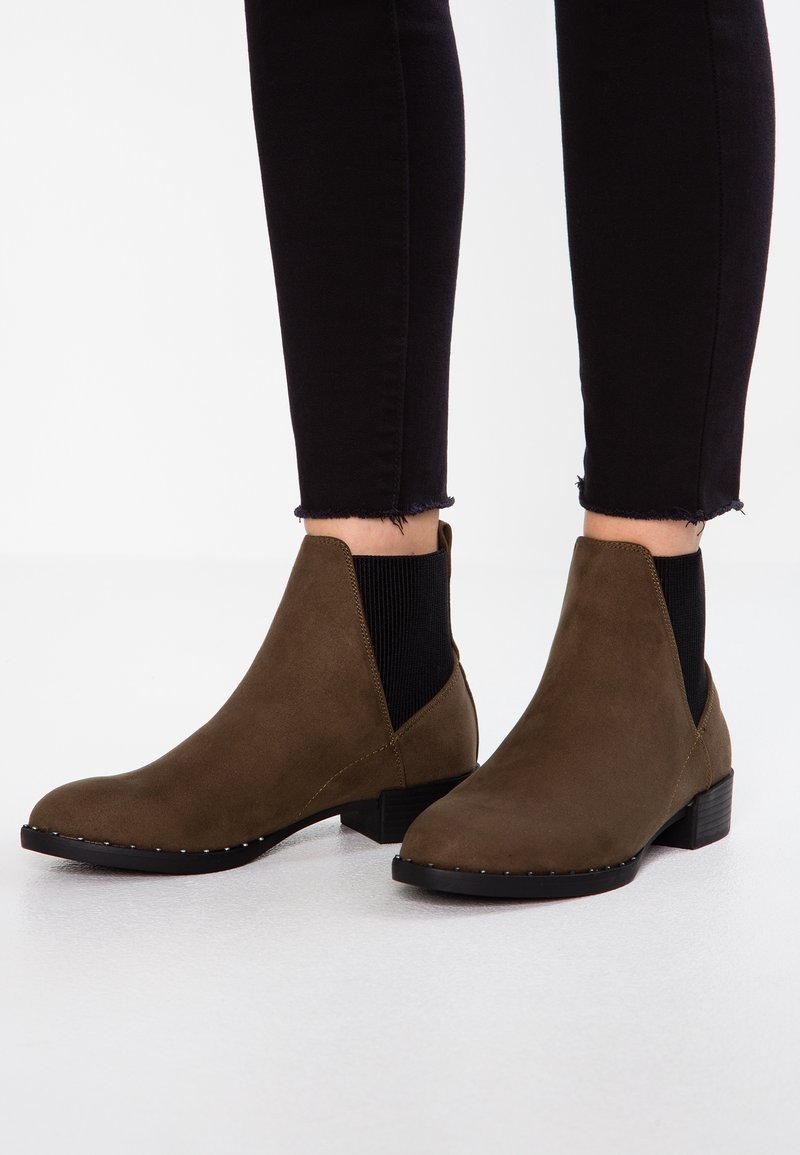 Personne portant des bottines en daim vert olive avec des côtés élastiques noirs et de petits talons carrés, associées à un jean skinny noir.