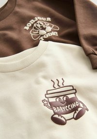 Primer plano de sudaderas marrones y crema con diseños de dibujos animados: un croissant con "Bonjour Today" y una taza de café sonriente etiquetada como "Babyccino".