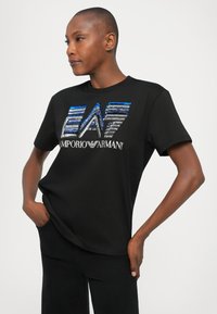 Cilvēks ar īsiem matiem, ģērbies melnā T-kreklā ar zilo un sudraba sekvīnu "EA7 Emporio Armani" logotipu un melnās biksēs, rokas kabatās.