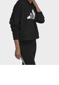 Felpa nera corta con grande logo bianco sul davanti, dotata di cappuccio e maniche lunghe e larghe. Abbinata a pantaloni neri.