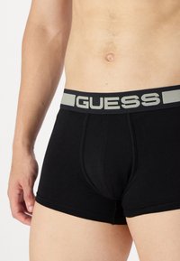 Juodi medvilniniai bokseriai su pilku elastingu juosmeniu, ant kurio yra "GUESS" logotipas. Lygus paviršius ir kūną apimantis dizainas.