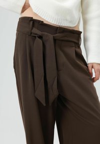 Bruine broek met hoge taille, met een strikceintuur, geplooide voorkant en zijzakken, gecombineerd met een witte gebreide trui.