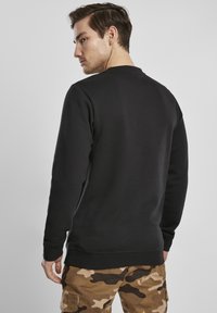 Melna džemperis ar apaļu apkakli un strukturētu apdari. Ribas malām uz aprocēm un apakšmalā. Modelis valkā kamuflāžas raksta bikses, radot ikdienišķu izskatu.