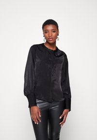 Blouse noire en satin à manches longues avec un col rond, une fermeture à boutons à l'avant et des poignets froncés. Texture lisse avec un léger éclat.