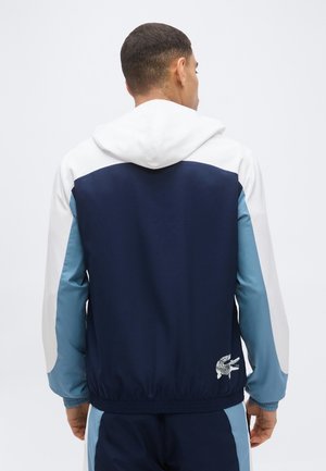 Hombre de espaldas con una chaqueta con capucha azul marino, blanca y azul claro y pantalones a juego con un pequeño logo blanco de cocodrilo cerca de la cintura.
