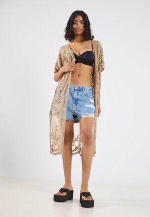 Beiger Spitzenkimono über einem schwarzen BH, kombiniert mit zerrissenen Jeansshorts. Schwarze Plateausandalen runden das Outfit ab.