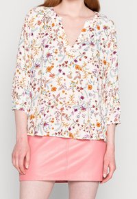 Weiße Bluse mit buntem Blumenmuster, mit einem V-Ausschnitt und Dreiviertelärmeln, kombiniert mit einem rosa Leder-Minirock.