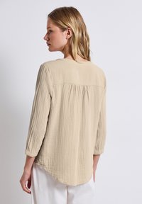 Helles beige Bluse aus einem strukturierten, zerknitterten Stoff, mit einem runden Ausschnitt und gerafftem Rückendetail. Lange Ärmel mit umgeschlagenen Bündchen.