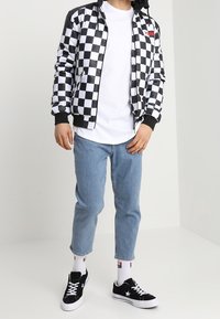 Giubbotto bomber a scacchi bianco e nero, t-shirt bianca, jeans azzurri chiari, sneakers nere e calze bianche con logo.