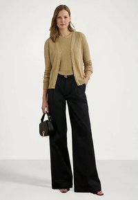 Γυναίκα φορώντας μπεζ ριγωτό κάρντιγκαν και πουλόβερ, μαύρο παντελόνι σε φαρδιά γραμμή, κρατώντας μαύρη τσάντα, στέκεται μπροστά σε απλό φωτεινό φόντο.